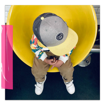 Un petit garçon de 3 ans assis sur un siège jaune, portant une casquette cool avec visière plate et un sweat coloré, style streetwear