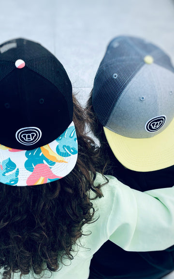 Deux enfants, une fille et un garçon, vus de dos assis dans un skatepark, portant des casquettes cool pour enfants style streetwear avec visière plate, noire avec motif imprimé tropical, grise avec visière jaune