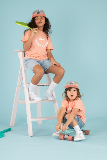 Enfants fille et garçon look streetwear sur un skate avec t-shirts cool pour enfants, coupe oversize unisexe en coton bio couleur pêche motif San Francisco, casquette snapback visière plate motif imprimé, short large en jean, baskets montantes, look matchy assorti frère soeur