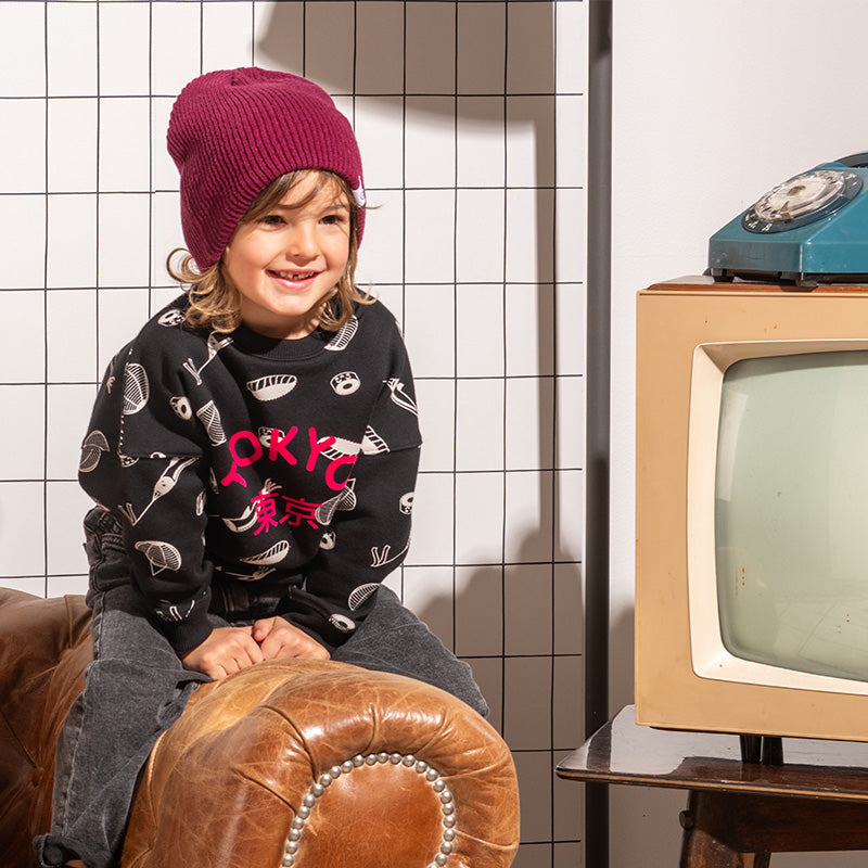 Petit garçon souriant look streetwear cool avec sweat unisexe coupe oversize en coton bio, motif imprimé Tokyo sushi style manga kawaii, bonnet pour enfant en laine couleur prune, jean baggy et baskets, décor salon vintage rétro