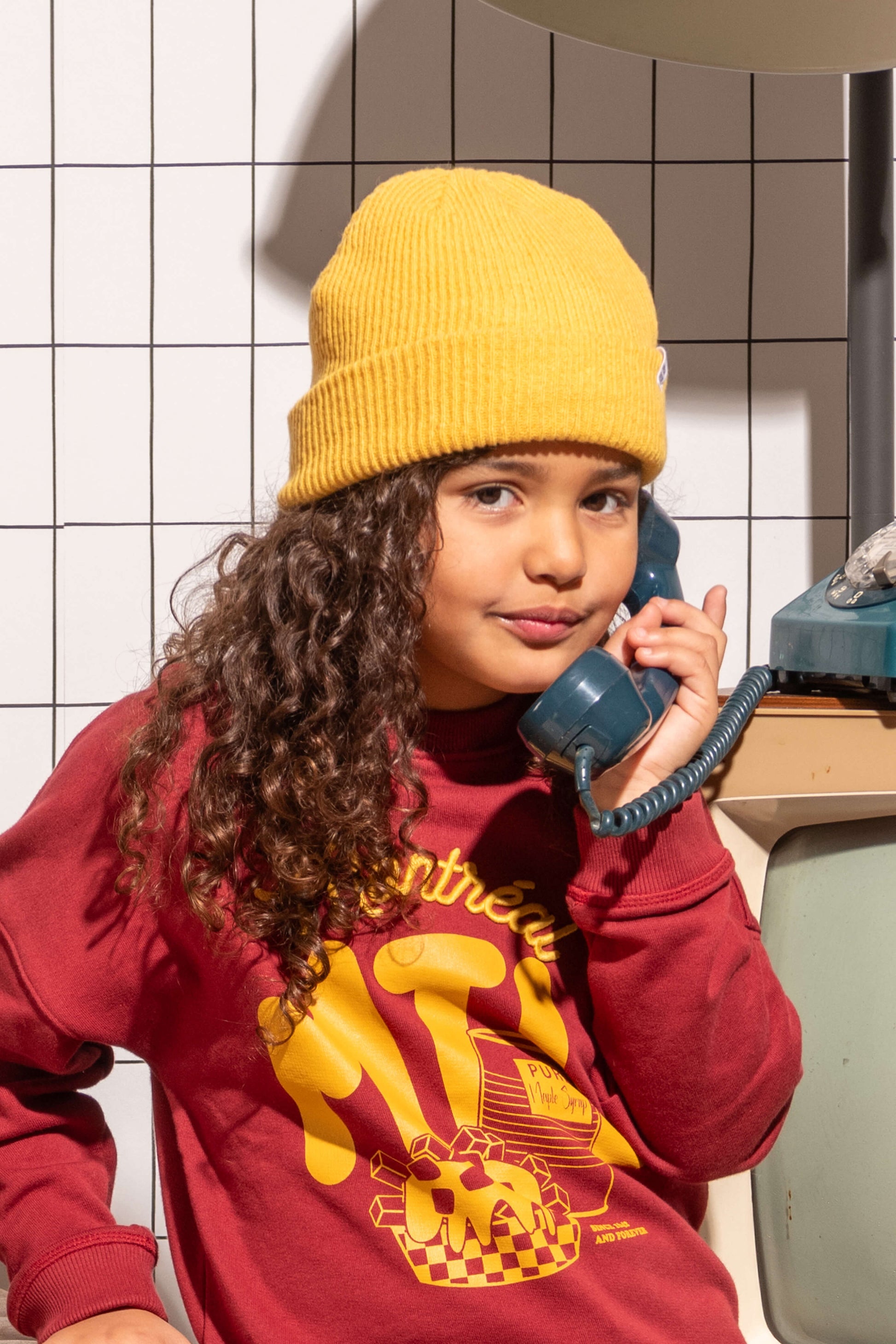 Fille look streetwear cool avec sweat pour enfant, unisexe, en coton bio, motif imprimé Montréal sur fond bordeaux, broderie en relief, bonnet pour enfant jaune moutarde, décor salon vintage avec téléphone rétro
