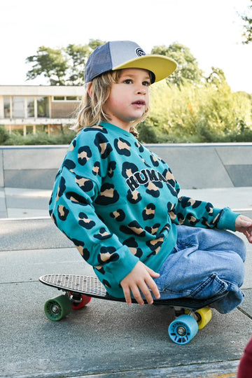 petit garçon dans un skatepark look streetwear cool sur un skate avec un sweat oversize unisexe en coton bio motif léopard bleu vert et une casquette snapback pour enfant visière plate grise et jaune