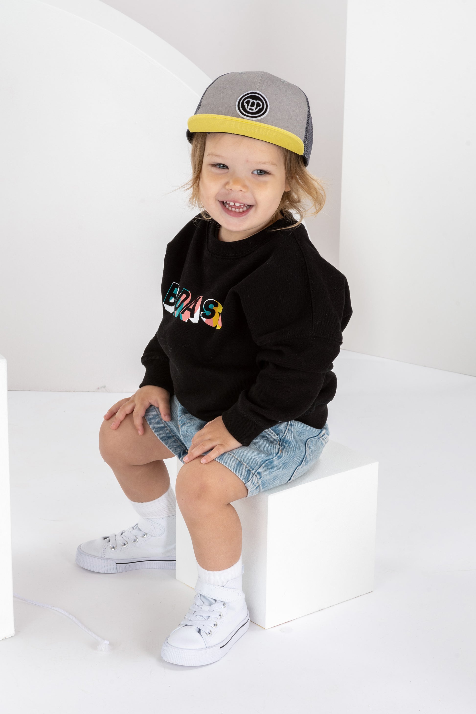 petite fille de 2 ans assise dans un décor monochrome blanc, en tenue streetwear cool, sweatshirt noir pour enfant imprimé Badass en coton bio, coupe unisexe oversize, casquette réglable pour enfant, visière plate grise et jaune, short en jean, baskets montantes blanches