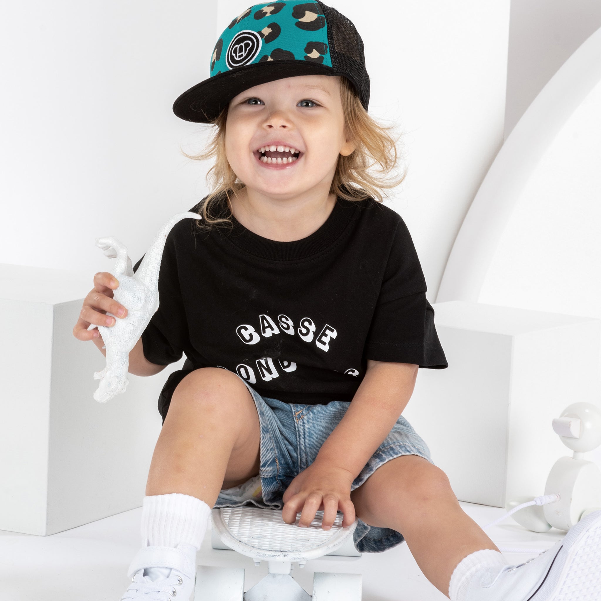 petite fille souriante de 2 ans, assise sur un skateboard, portant une tenue streetwear cool, casquette pour enfant, réglable, en coton et maille filet, visière plate, motif imprimé léopard bleu vert, visière noire, logo ours Allmemberz brodé devant, t-shirt noir en coton bio imprimé casse bonbons