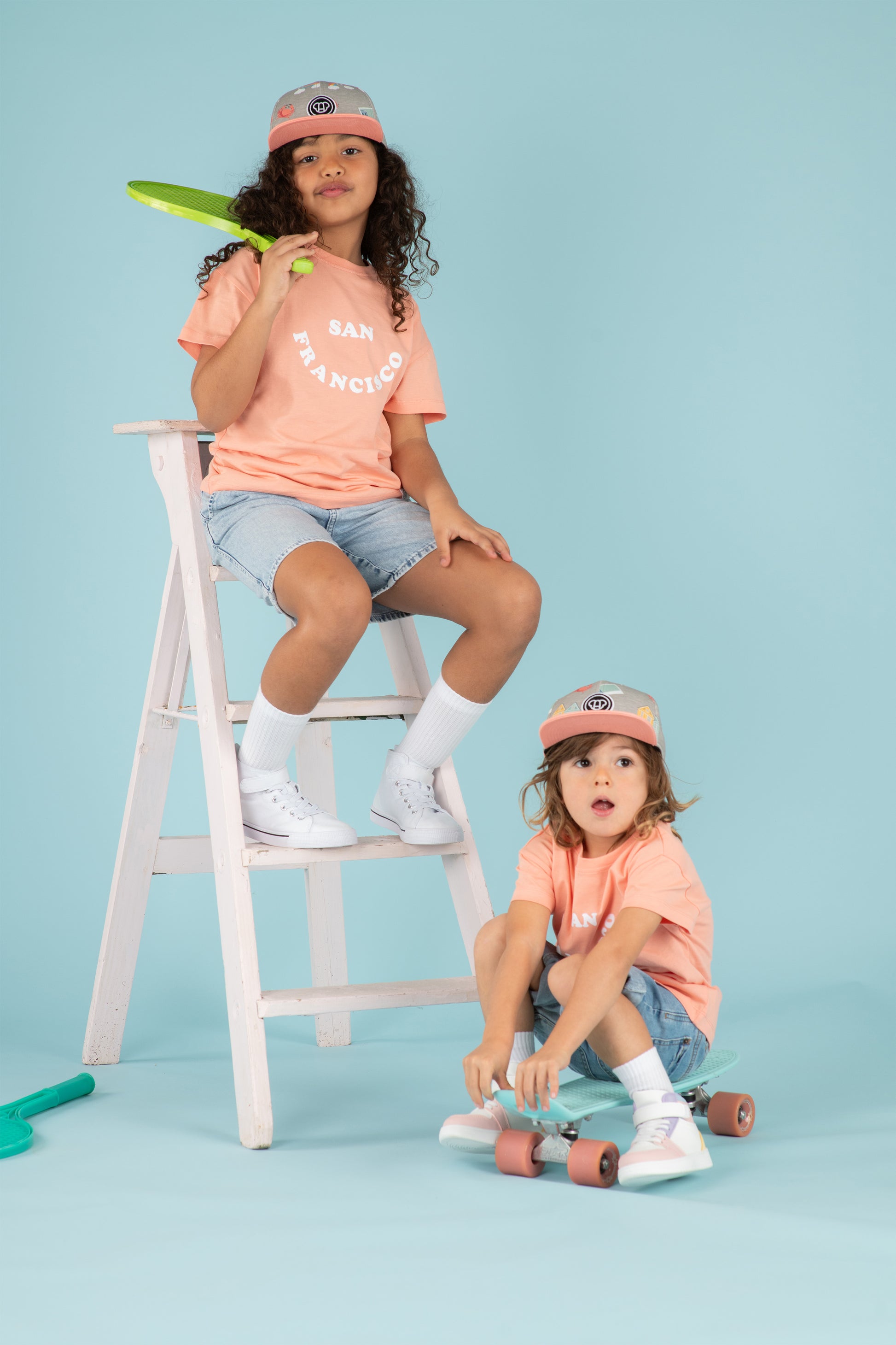 Enfants fille et garçon look streetwear sur un skate avec t-shirts cool pour enfants, coupe oversize unisexe en coton bio couleur pêche motif San Francisco, casquette snapback visière plate motif imprimé, short large en jean, baskets montantes, look matchy assorti frère soeur