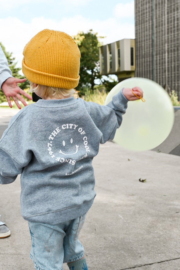 petit garçon dans un skatepark look streetwear cool, sweat oversize gris en coton bio motif happy face style retro au dos, bonnet pour enfant en laine jaune moutarde