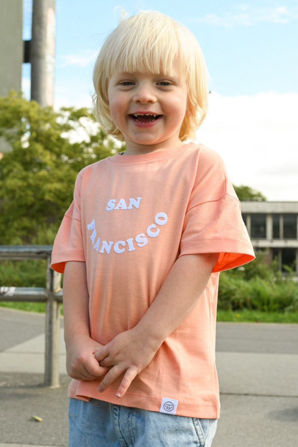 petit garçon souriant dans un skatepark, look streetwear cool avec t-shirt oversize unisexe en coton bio couleur pêche motif San Francisco