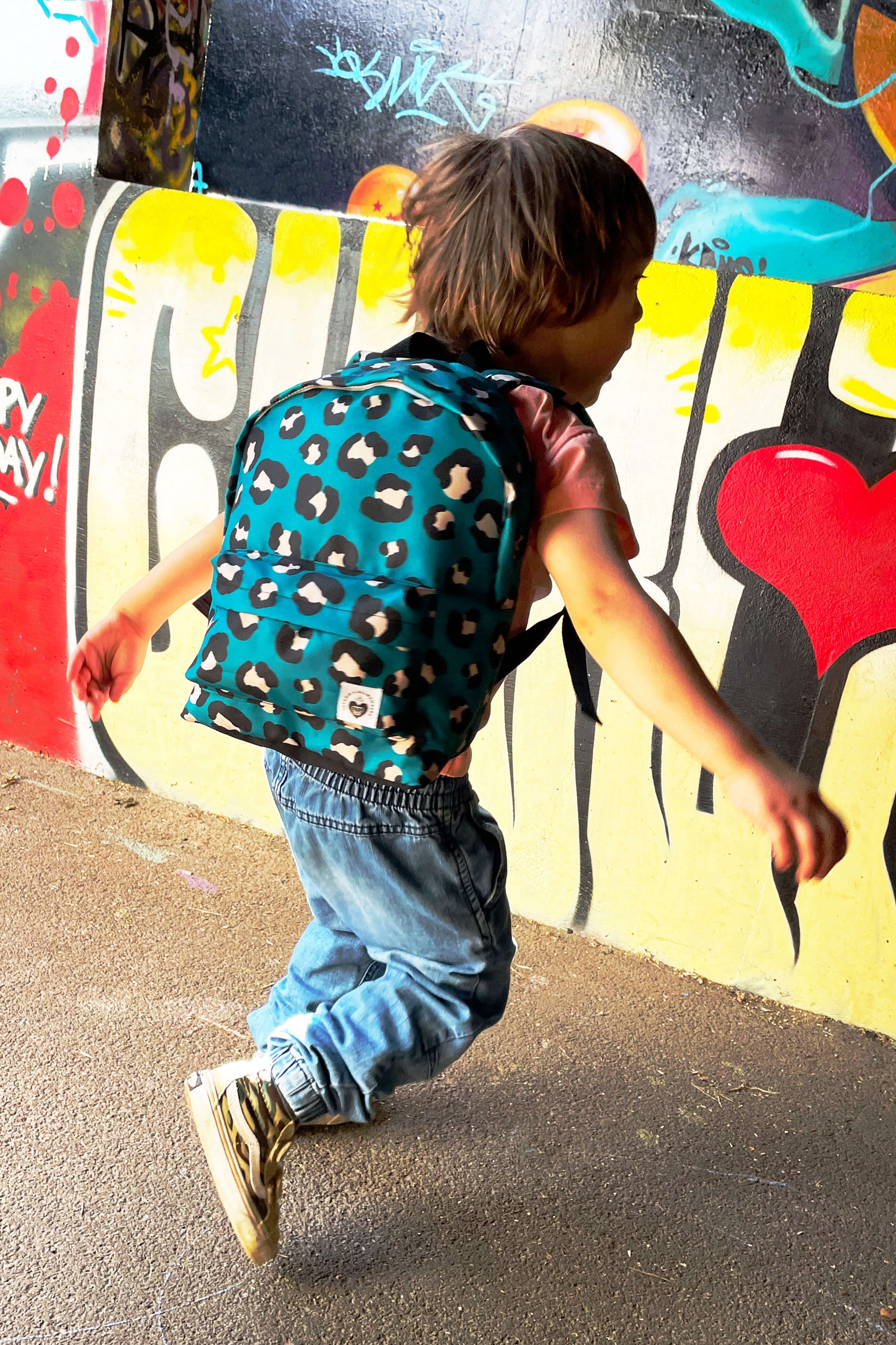 petit garçon look streetwear dans un décor urbain graffiti, portant un sac à dos pour enfant motif léopard fond bleu vert