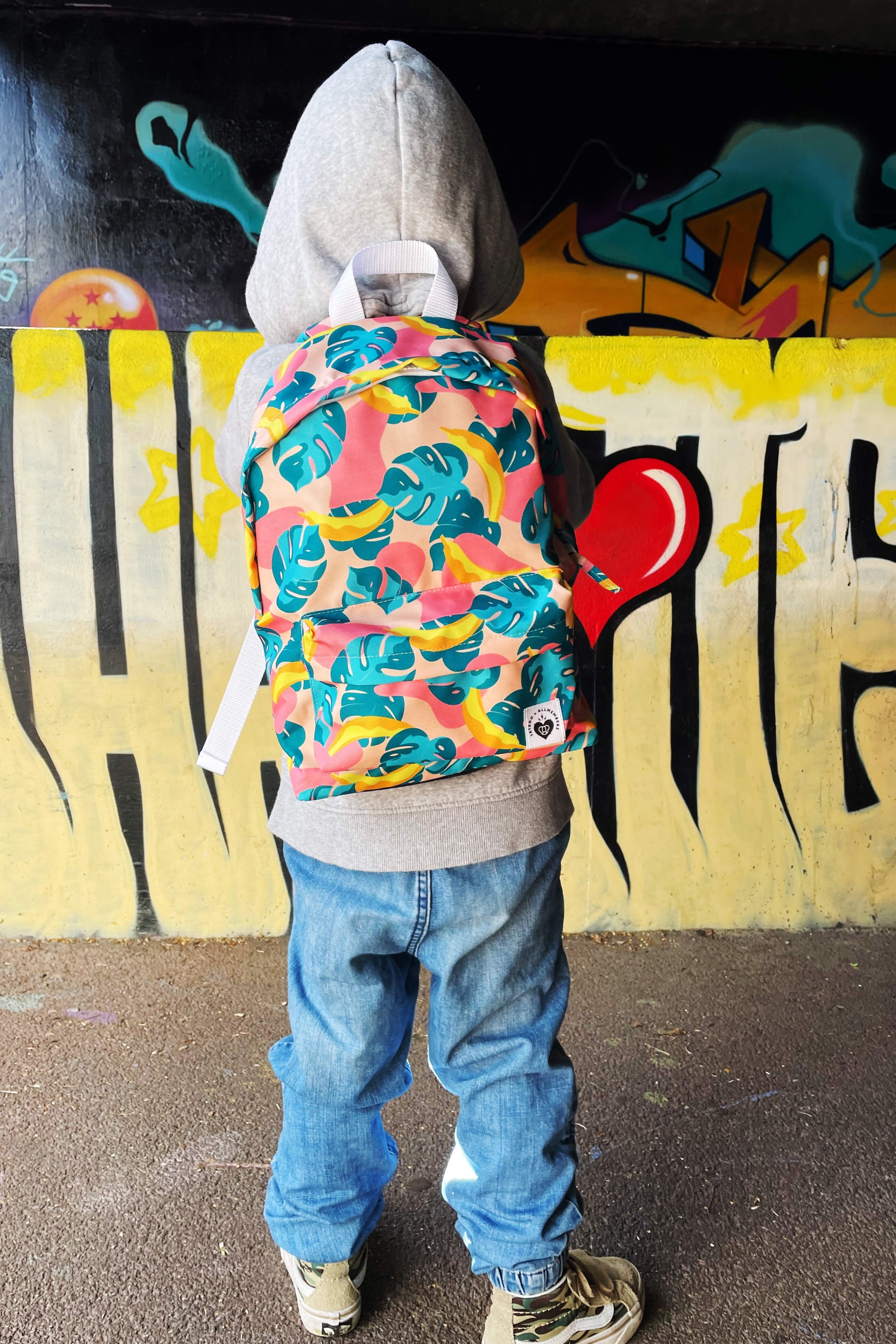 petit garçon look streetwear dans un décor urbain graffiti, portant un sac à dos pour enfant motif tropical joyeux et coloré, feuilles et bananes