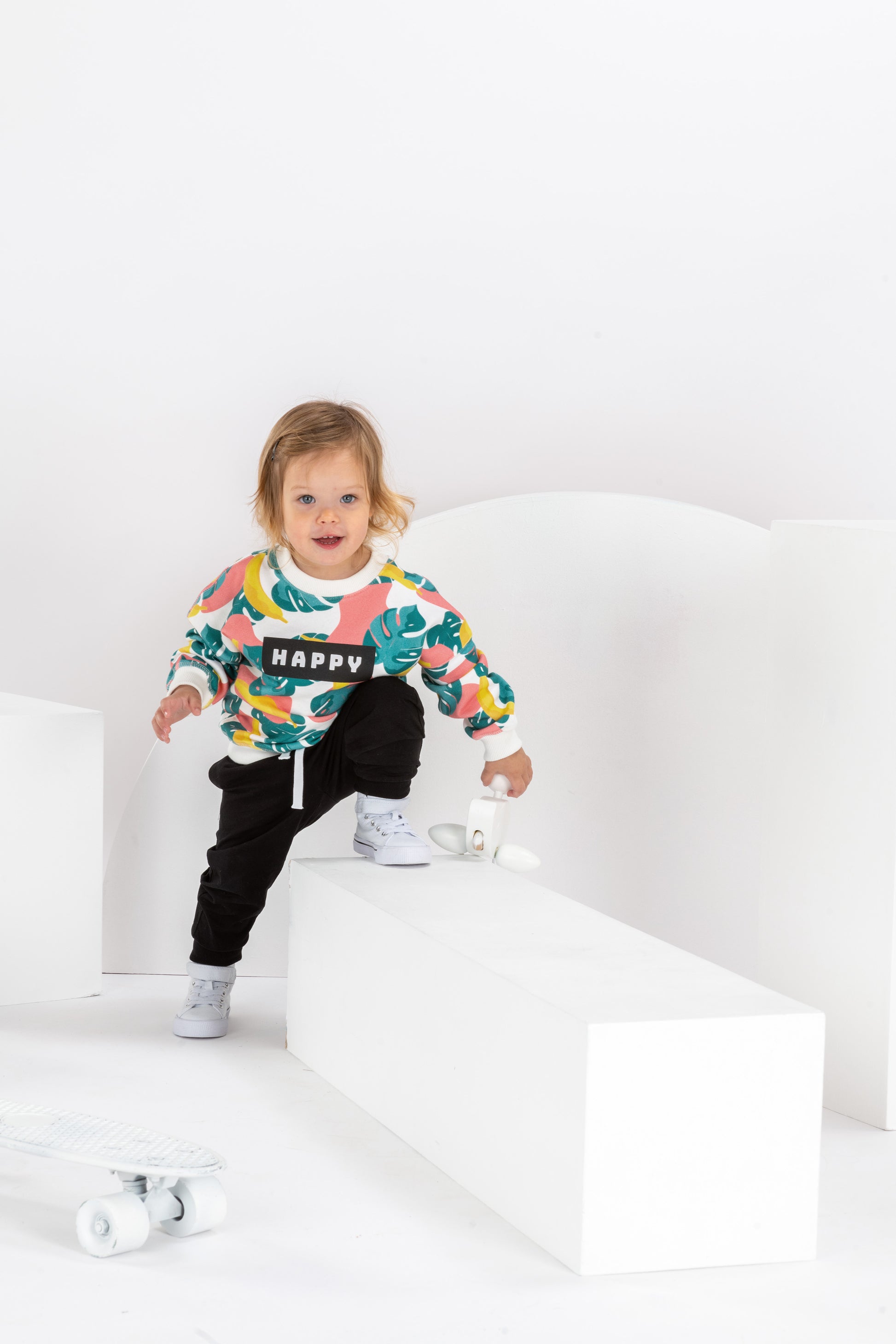 petite fille souriante de 2 ans, dans un décor monochrome blanc, en tenue streetwear cool avec sweat pour enfant, coupe unisexe oversize en coton bio, motif imprimé Happy tropical coloré, motif bananes, pantalon jogging noir et baskets montantes blanches