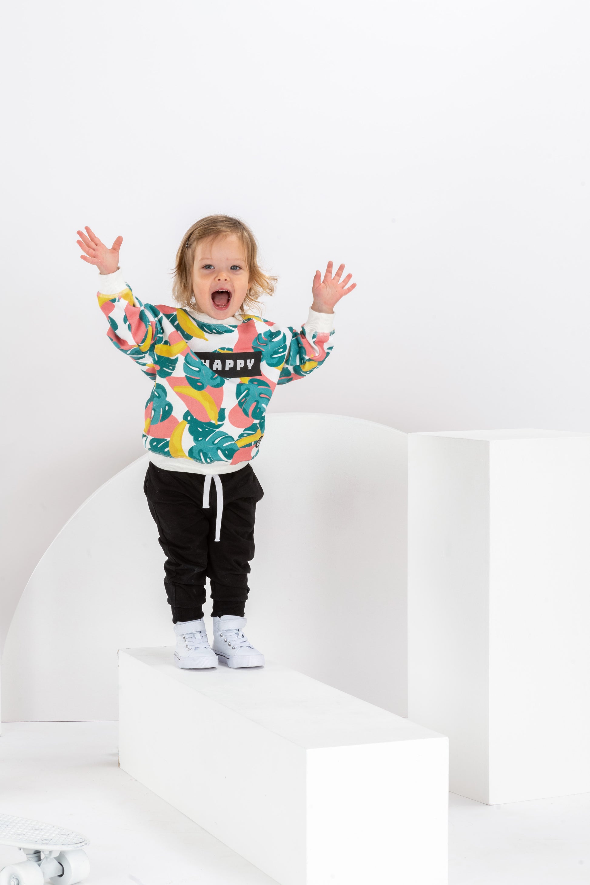 petite fille souriante de 2 ans, dans un décor monochrome blanc, en tenue streetwear cool avec sweat pour enfant, coupe unisexe oversize en coton bio, motif imprimé Happy tropical coloré, motif bananes, pantalon jogging noir et baskets montantes blanches
