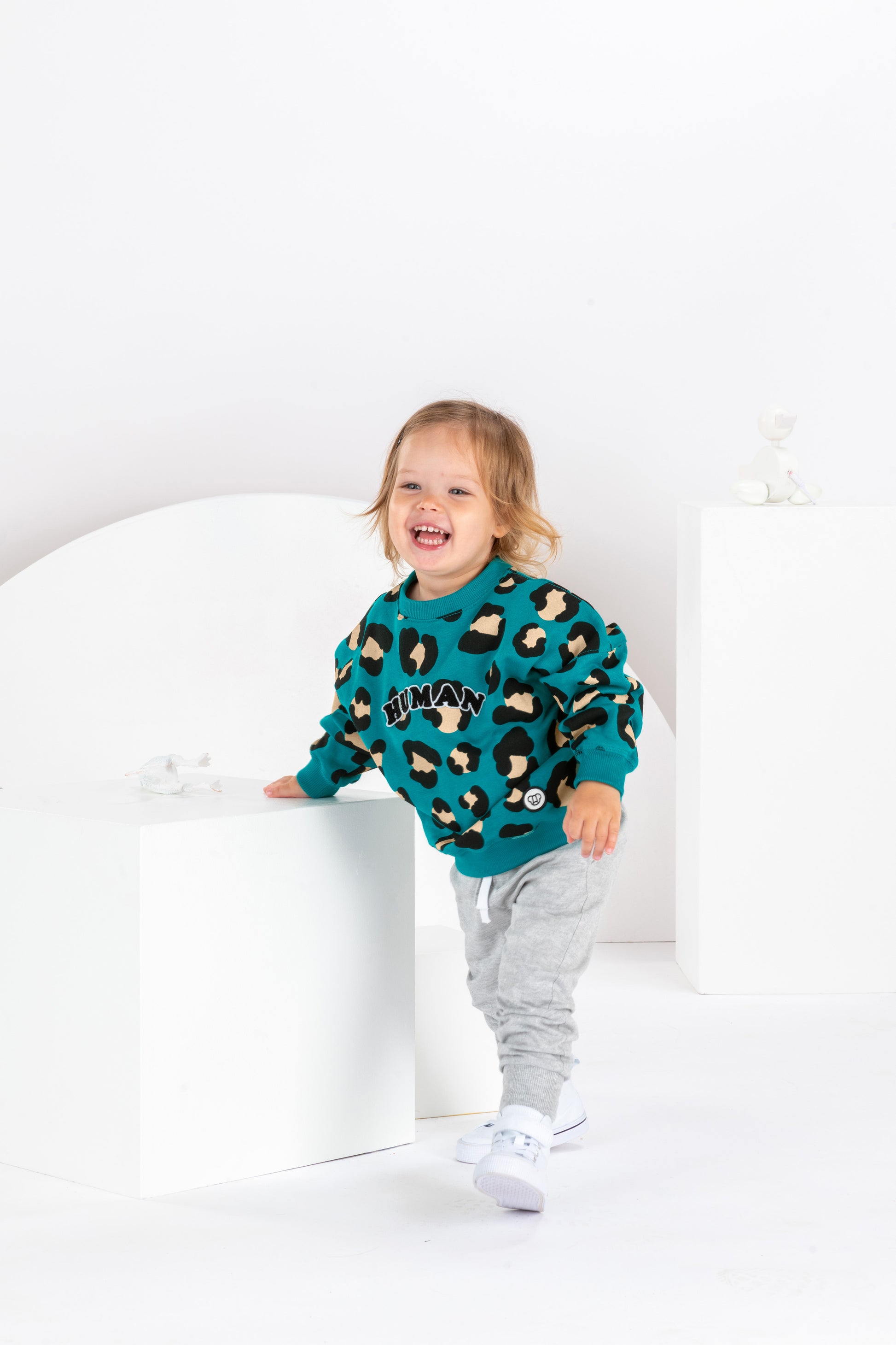 Petite fille de 2 ans souriante, sweat unisexe coupe oversize en coton bio, motif imprimé léopard bleu vert, look streetwear cool, baggy jogging gris, baskets montantes blanches, décor monocrome blanc