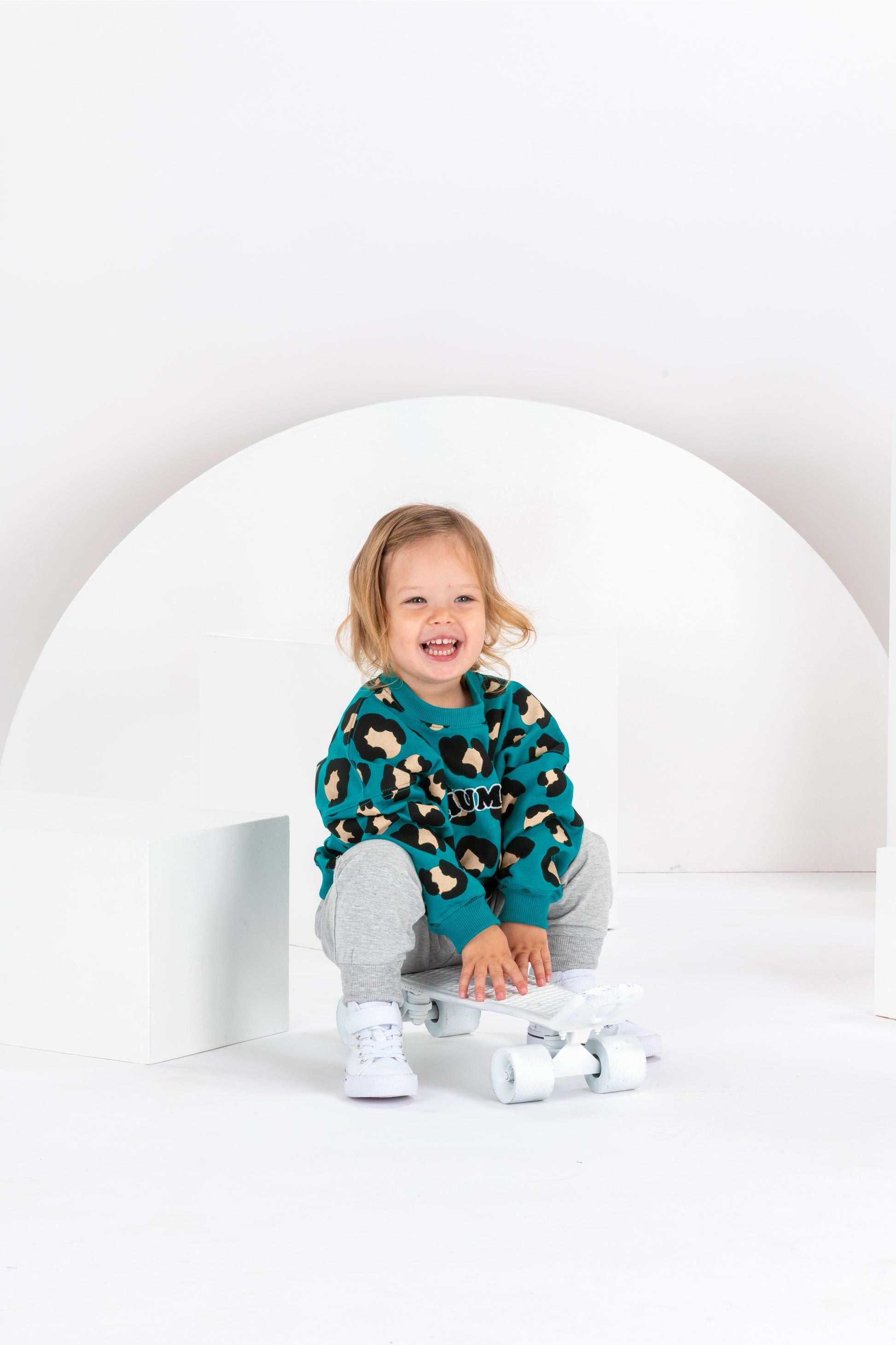Petite fille de 2 ans souriante assise sur un skateboard, sweat unisexe coupe oversize en coton bio, motif imprimé léopard bleu vert, look streetwear cool, baggy jogging gris, baskets montantes blanches, décor monocrome blanc