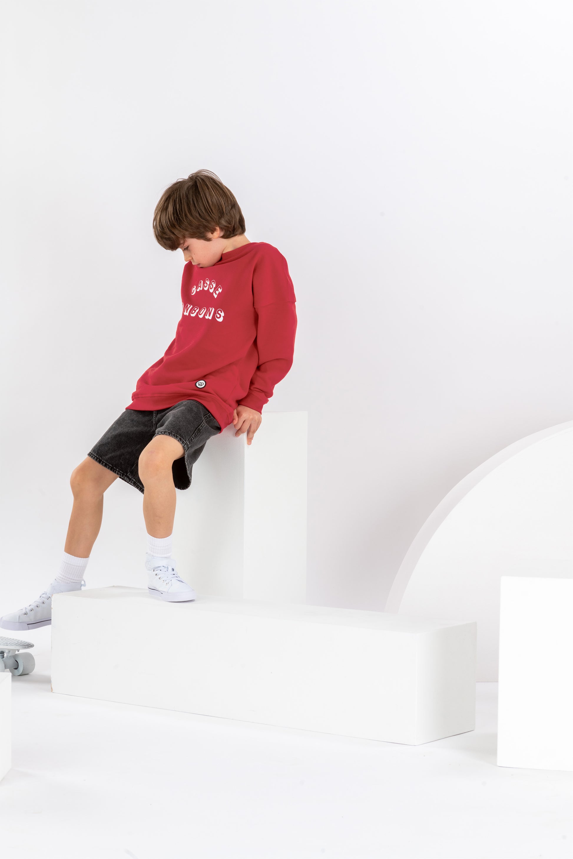 garçon de 8 ans dans un décor monochrome blanc, en tenue streetwear cool, sweat rouge pour enfant avec imprimé drôle Casse Bonbons, coupe unisexe oversize en coton bio, short en jean noir et baskets blanches montantes