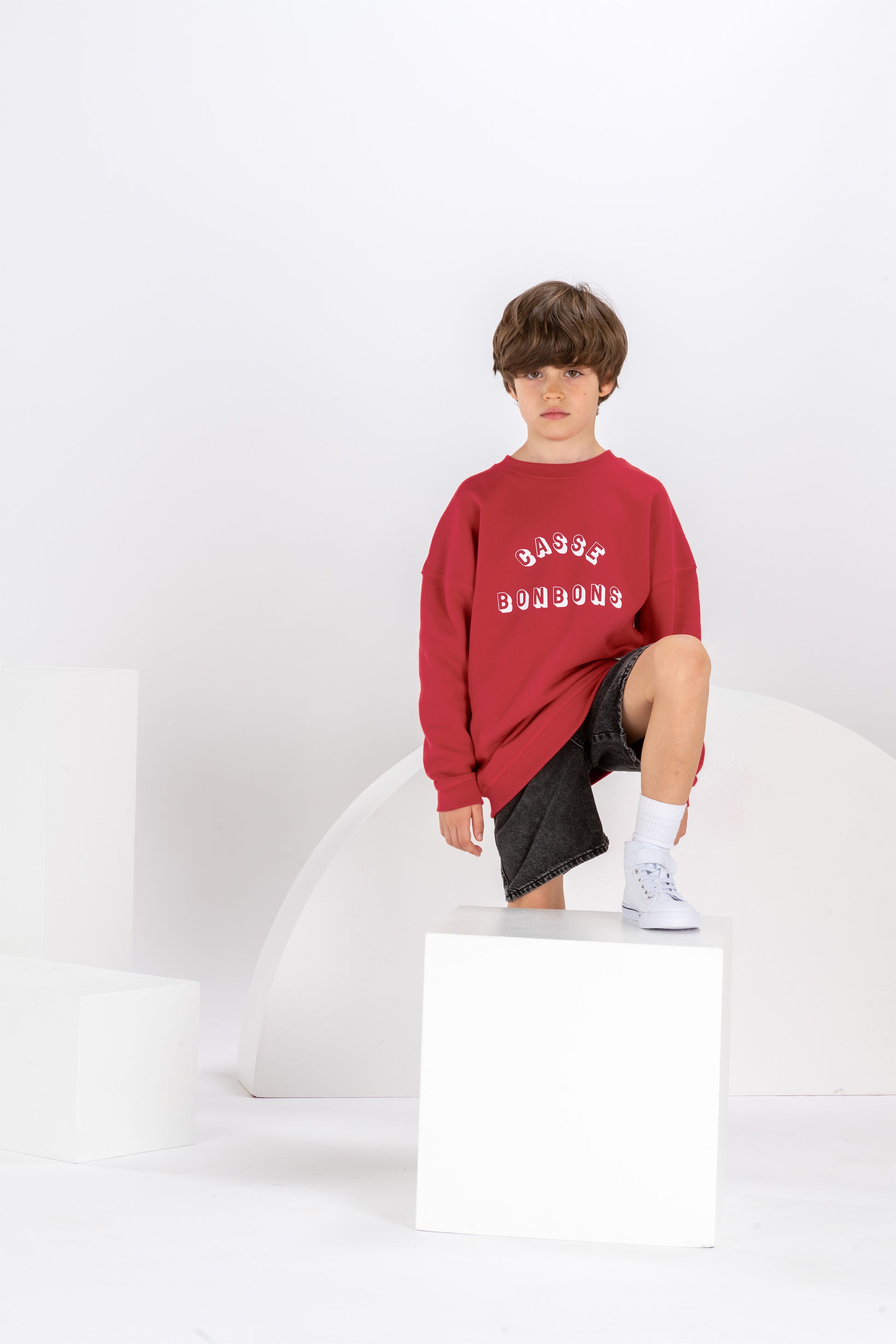 garçon de 8 ans dans un décor monochrome blanc, en tenue streetwear cool, sweat rouge pour enfant avec imprimé drôle Casse Bonbons, coupe unisexe oversize en coton bio, short en jean noir et baskets blanches montantes