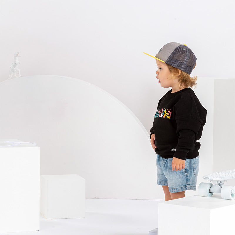 petite fille de 2 ans dans un décor monochrome blanc avec un skateboard, en tenue streetwear cool, sweatshirt noir pour enfant imprimé Badass en coton bio, coupe unisexe oversize, casquette réglable pour enfant, visière plate grise et jaune, short en jean