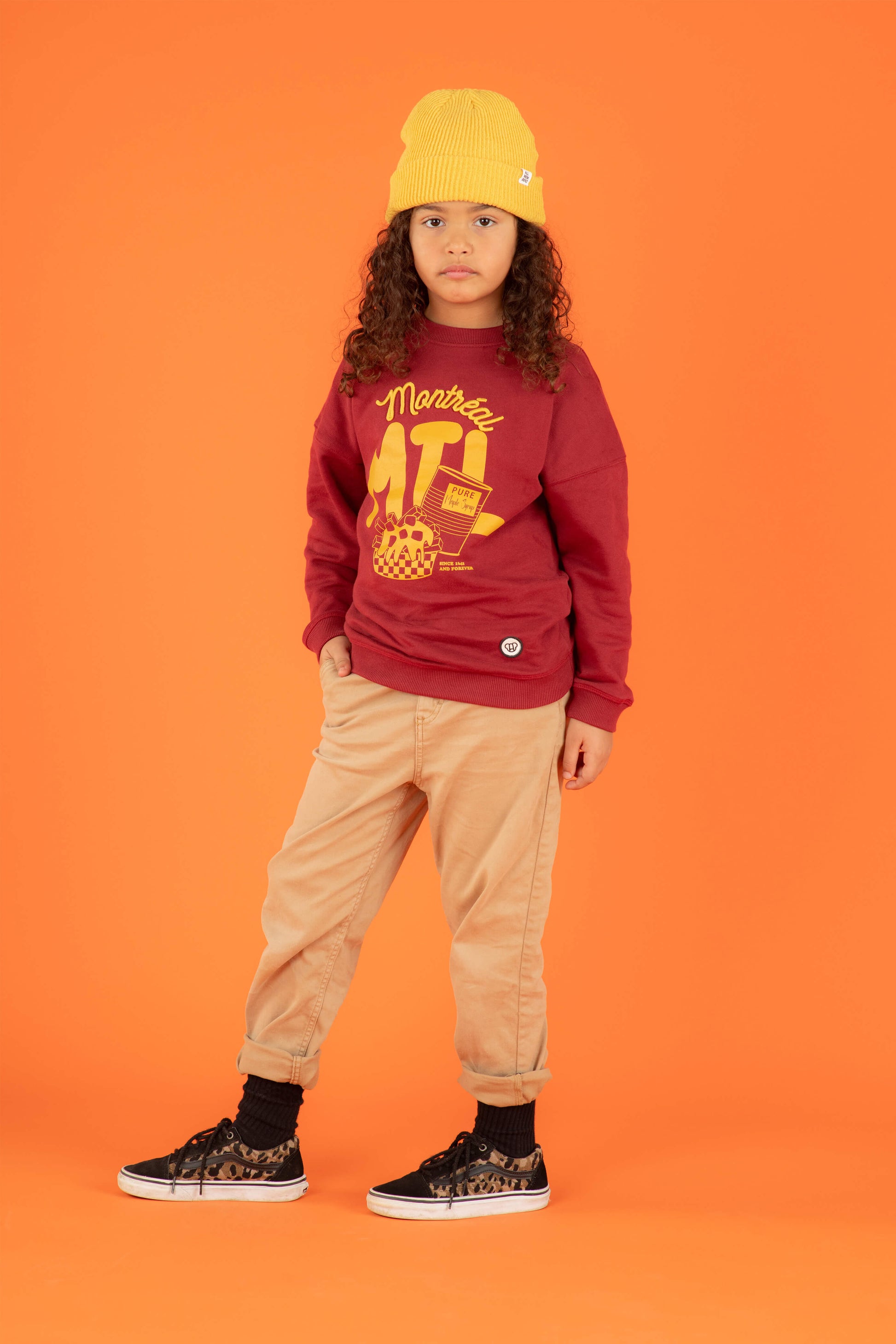 Fille look streetwear cool avec sweat unisexe bordeaux coupe oversize en coton bio, motif imprimé illustration Montréal, bonnet pour enfant en laine couleur jaune moutarde, jeans baggy, décor couleur orange