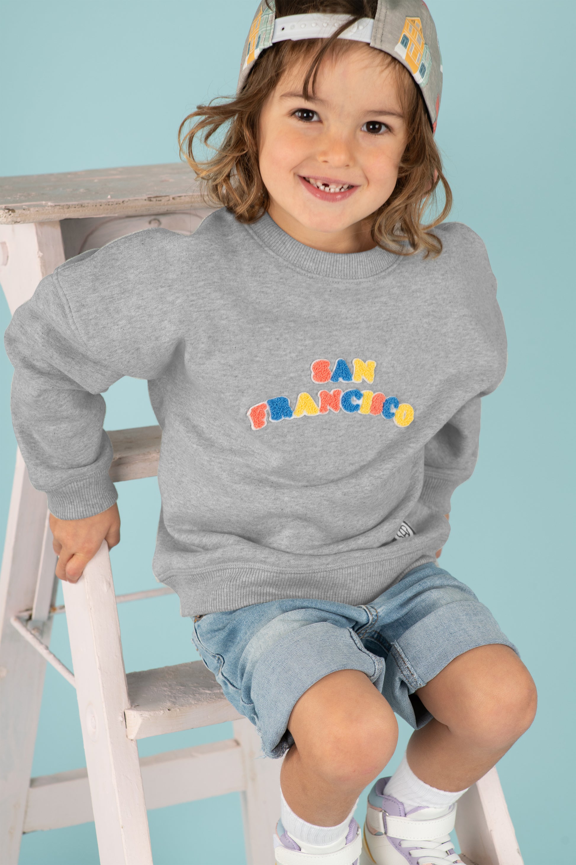 Petit garçon souriant look streetwear cool, sweat pour enfants coupe oversize unisexe gris chiné en coton bio motif multicolore San Francisco, casquette snapback enfant visière plate motif imprimé couleurs pastel, short large en jean