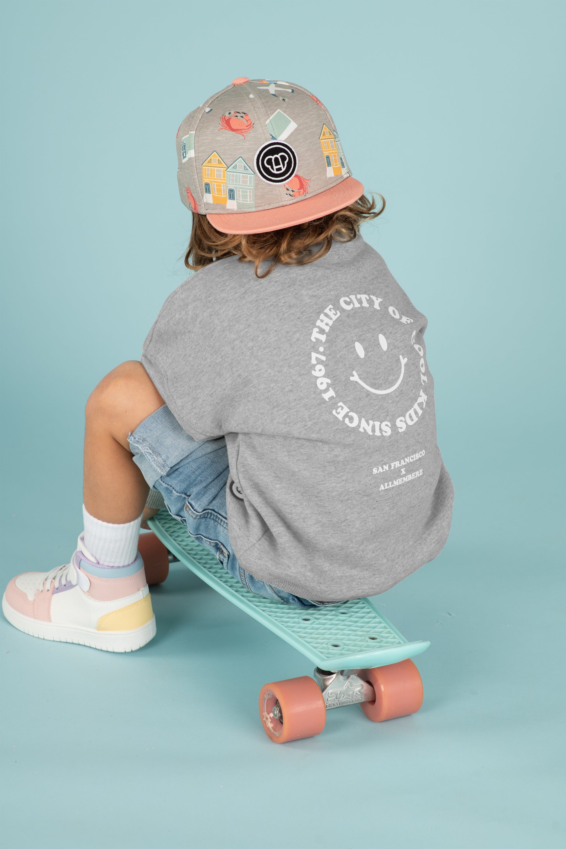 Petit garçon sur un skate look streetwear cool, sweat pour enfants coupe oversize unisexe gris chiné en coton bio motif imprimé happy face San Francisco, casquette snapback enfant visière plate motif imprimé couleurs pastel, baskets montantes multicolores