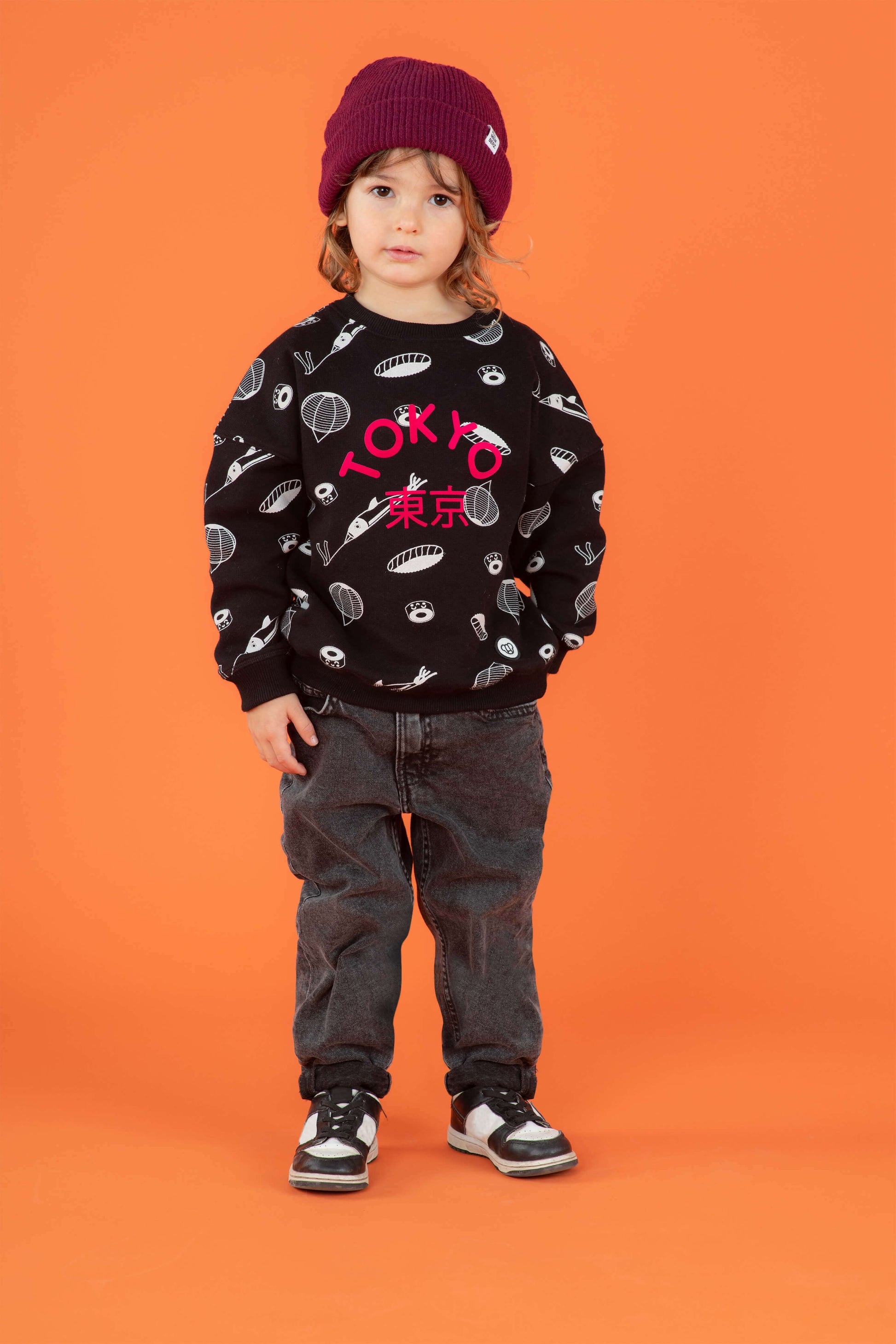 Petit garçon look streetwear cool avec sweat unisexe coupe oversize en coton bio, motif imprimé Tokyo sushi style manga kawaii, bonnet pour enfant en laine couleur prune, jean baggy et baskets, décor fond orange
