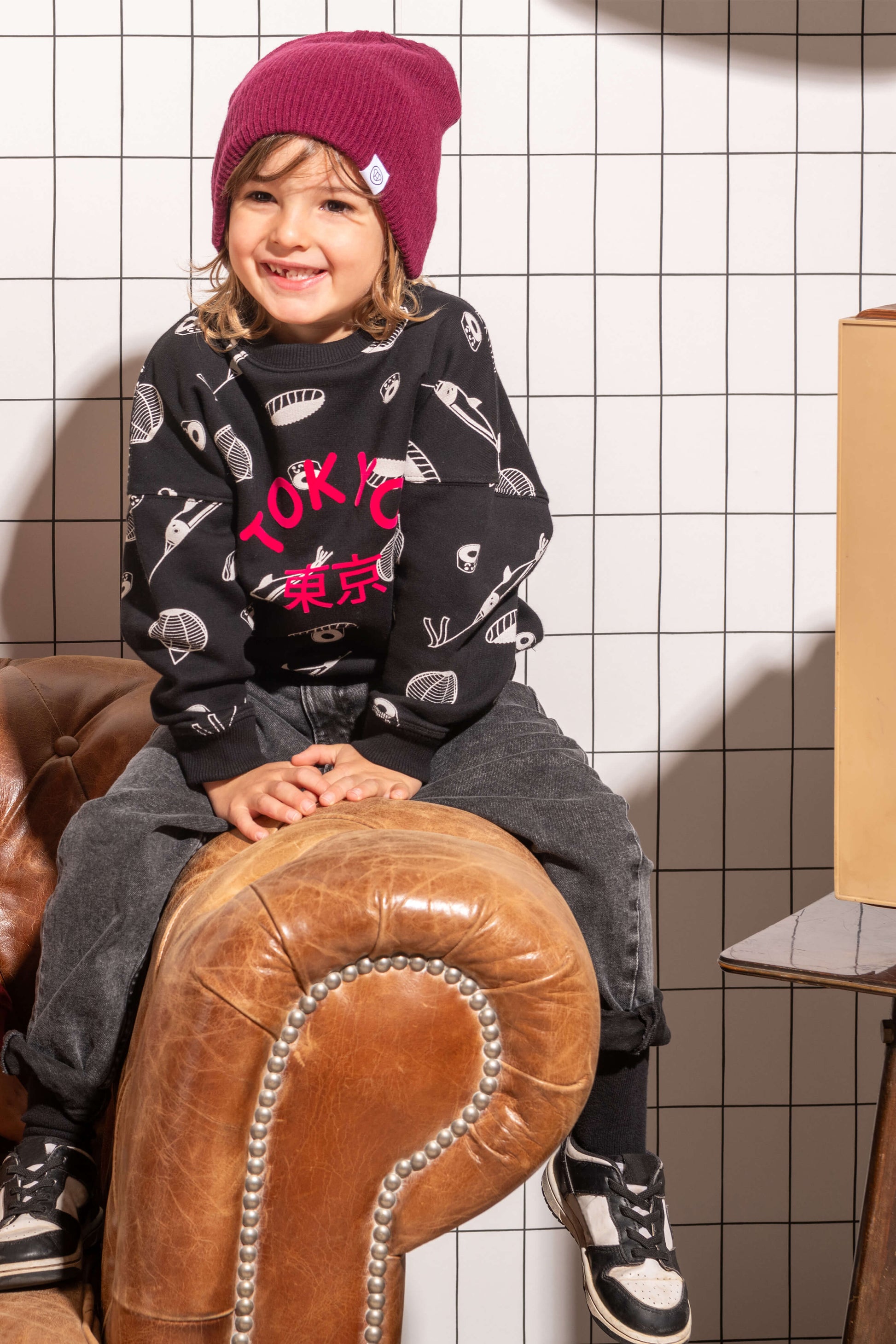 Petit garçon souriant look streetwear cool avec sweat unisexe coupe oversize en coton bio, motif imprimé Tokyo sushi style manga kawaii, bonnet pour enfant en laine couleur prune, jean baggy et baskets, décor salon vintage canapé cuir rétro