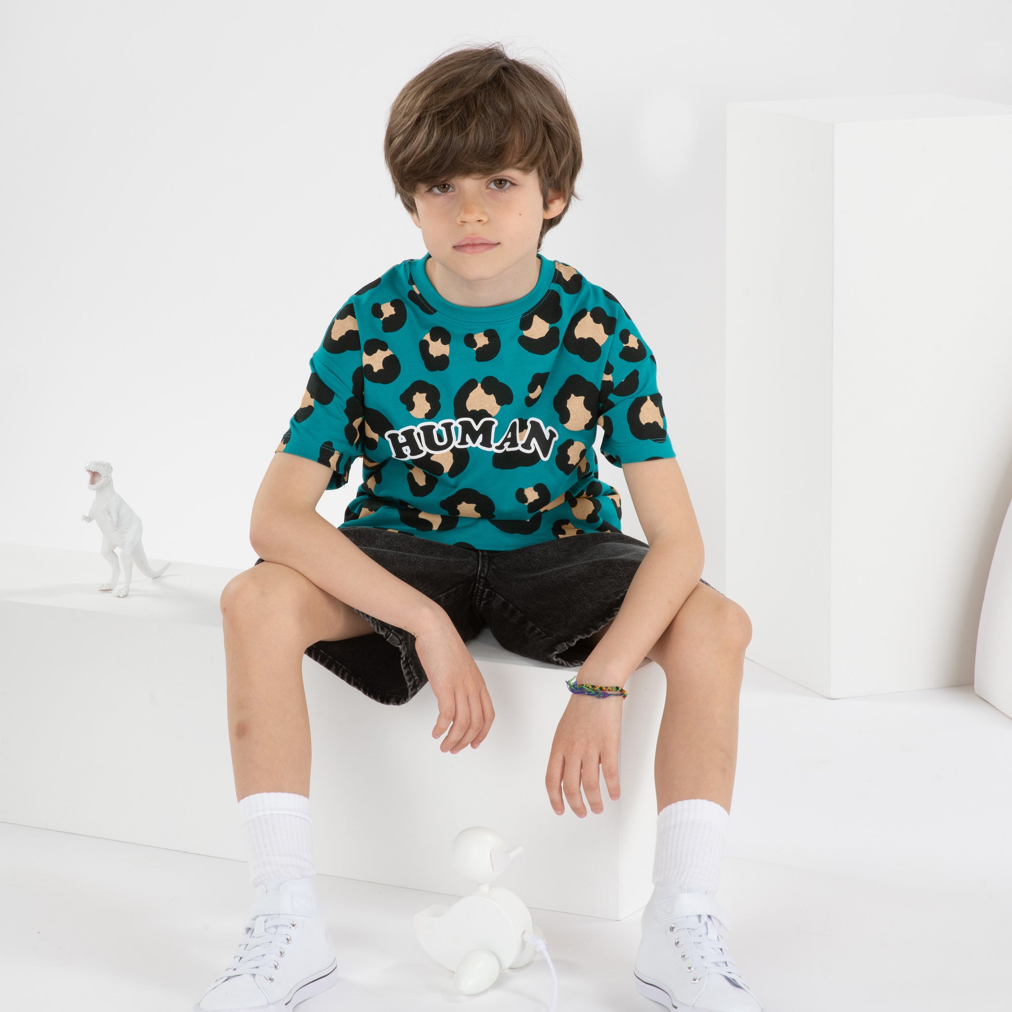 Garçon de 8 ans portant un t-shirt streetwear cool pour enfant, coupe oversize unisexe en coton biologique, motif imprimé léopard fond vert bleu, short large jean noir, baskets montantes blanches, dans un décor monochrome blanc
