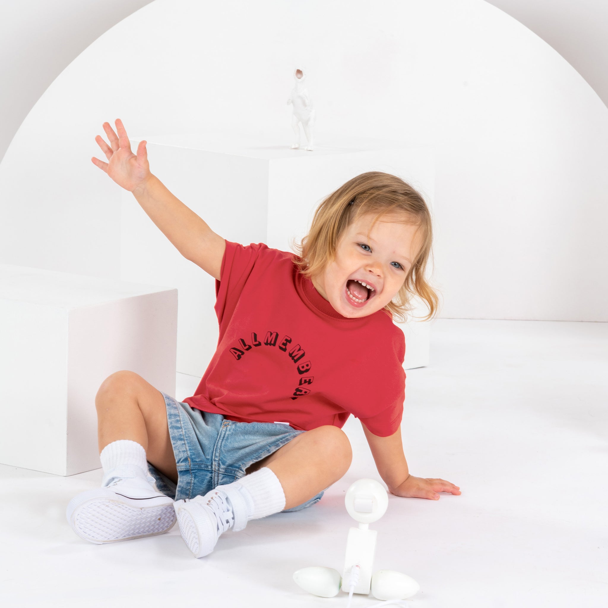 petite fille souriante de 2 ans, assise dans un décor monochrome blanc, en tenue streetwear avec t-shirt rouge cerise pour enfant, en coton bio, coupe oversize unisexe, motif imprimé Allmemberz, short en jean, baskets montantes blanches