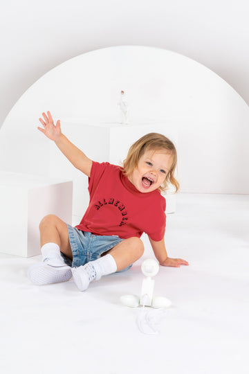 petite fille souriante de 2 ans, assise dans un décor monochrome blanc, en tenue streetwear avec t-shirt rouge cerise pour enfant, en coton bio, coupe oversize unisexe, motif imprimé Allmemberz, short en jean, baskets montantes blanches